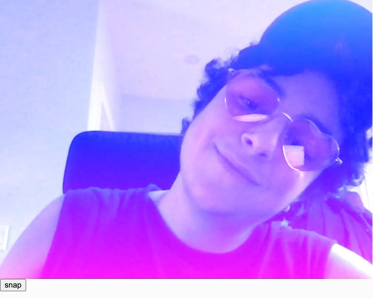 A white person with short dark curly hair, a dark sleeveless t-shirt, and pink heart shaped glasses tilts his head and looks down at the camera, smiling softly. A blue to pink vertical linear gradient is overlaid on top of the photo. Because of the LIGHTEST blendMode we're using, the colorful gradient is especially visible on his dark hair and dark shirt.
